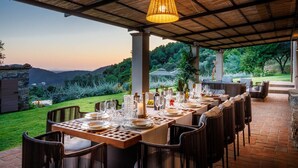 Un espace de restauration en plein air avec une longue table dressée pour un repas, entourée de chaises en osier, sous une pergola en bois agrémentée d’un luminaire suspendu.