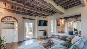 Un salon spacieux avec un plafond haut, des poutres apparentes, un grand canapé et un téléviseur à écran plat fixé au mur.