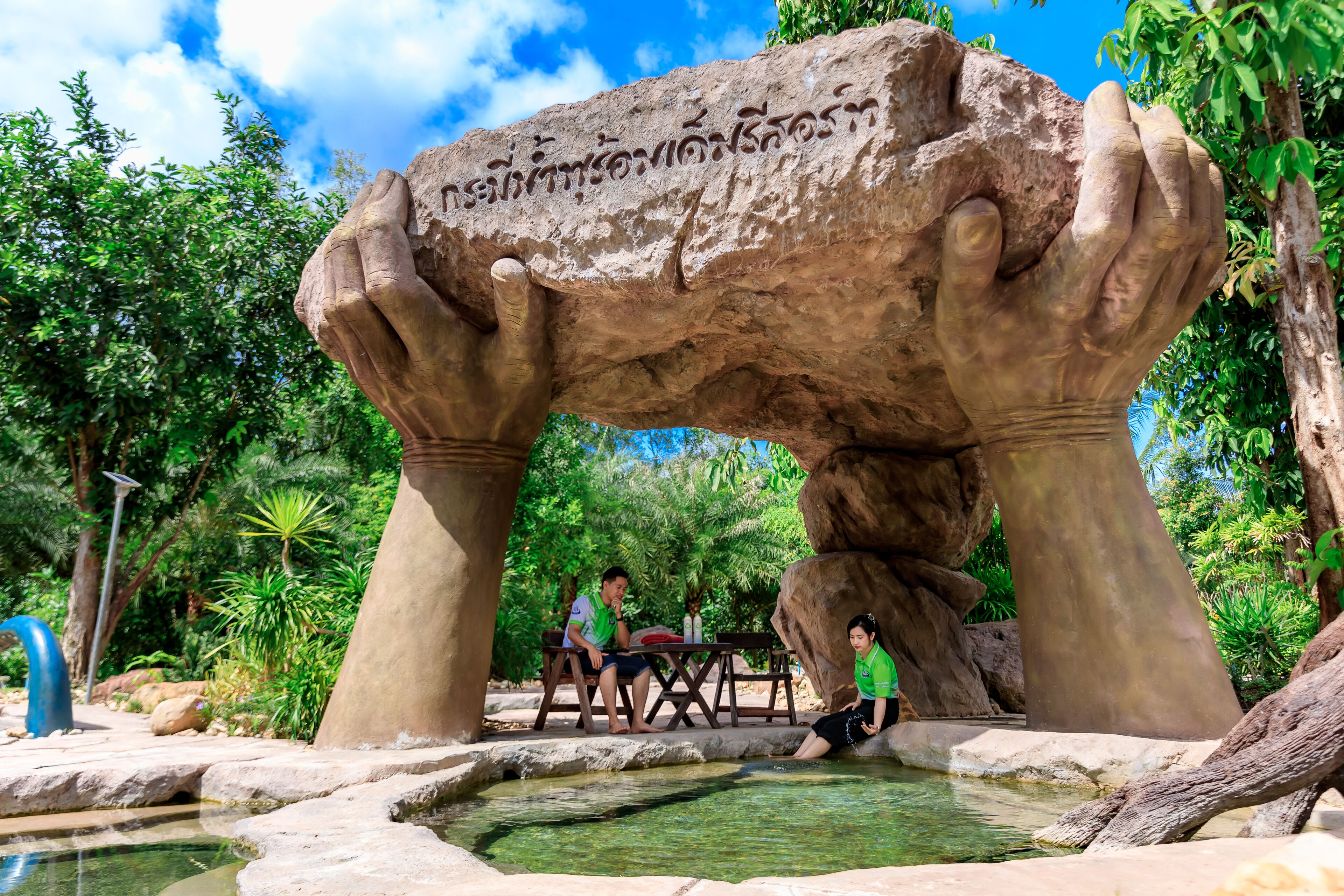 A rock archway with inscriptions, a small pool, and people sitting under it.