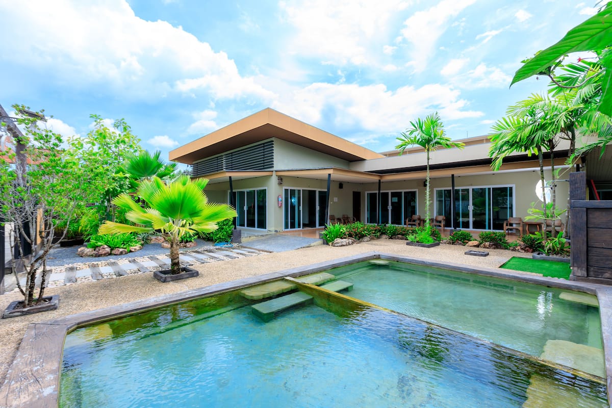 Ein modernes Haus mit Swimmingpool, umgeben von tropischen Pflanzen und blauem Himmel.