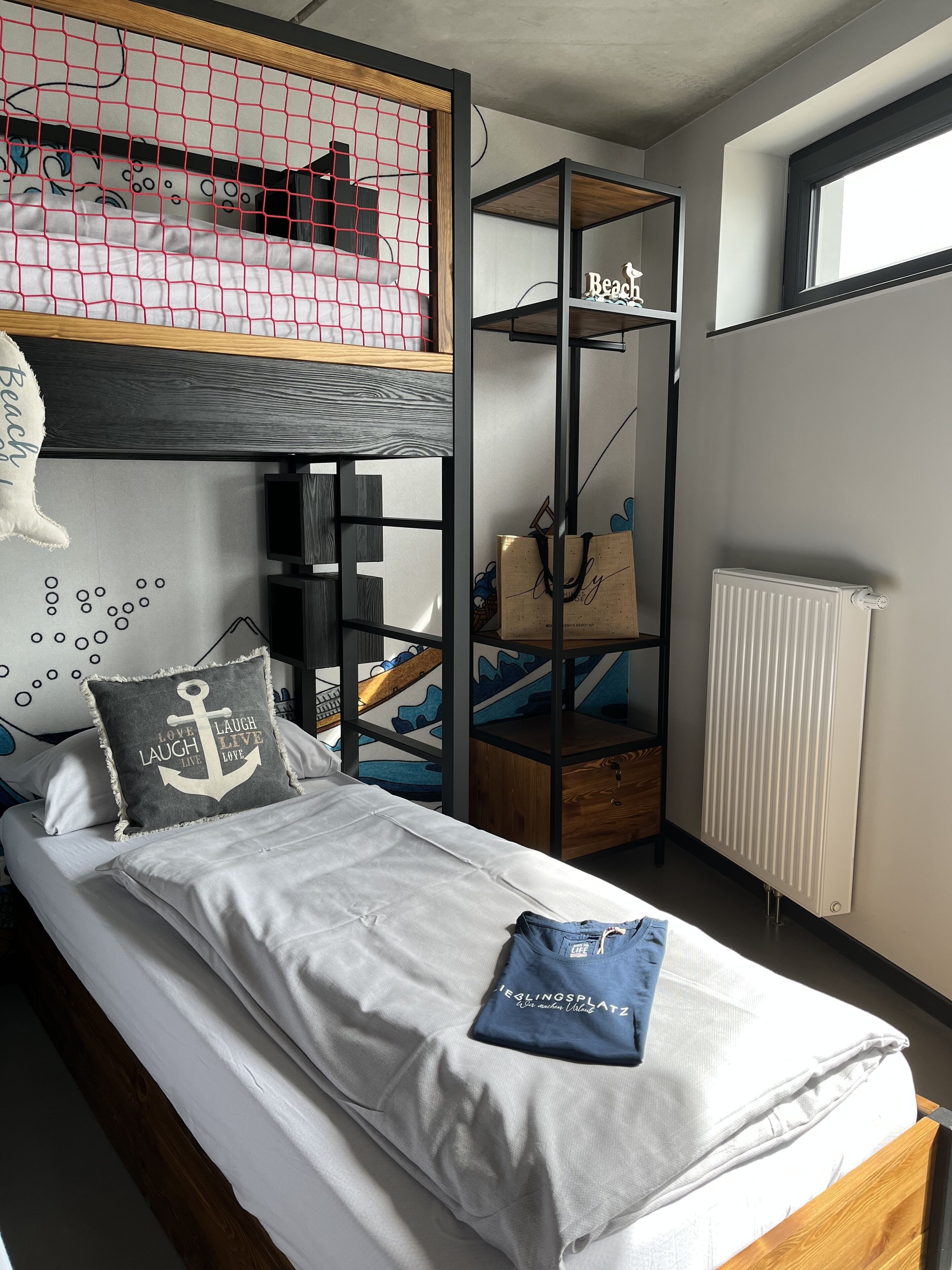Ein Etagenbett mit Moskitonetz im oberen Bereich, ein Nachttisch mit Lampe, eine Heizung und ein Regal mit einer Tasche und einem Schild mit der Aufschrift 'Beach'.