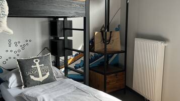 Ein Etagenbett mit Moskitonetz im oberen Bereich, ein Nachttisch mit Lampe, eine Heizung und ein Regal mit einer Tasche und einem Schild mit der Aufschrift 'Beach'.