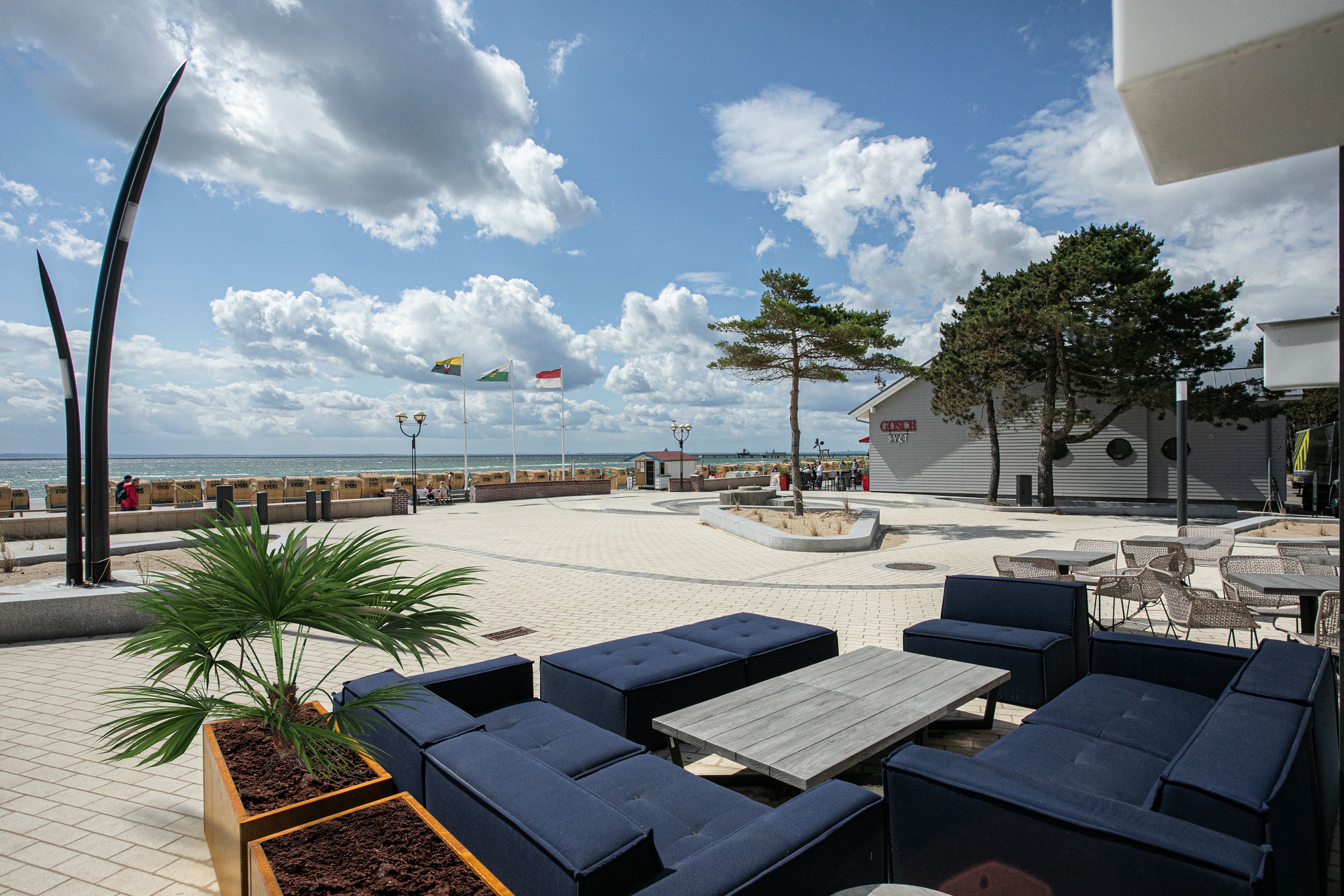An outdoor seating area with lounge chairs and tables, a potted plant, and a view of the sea.