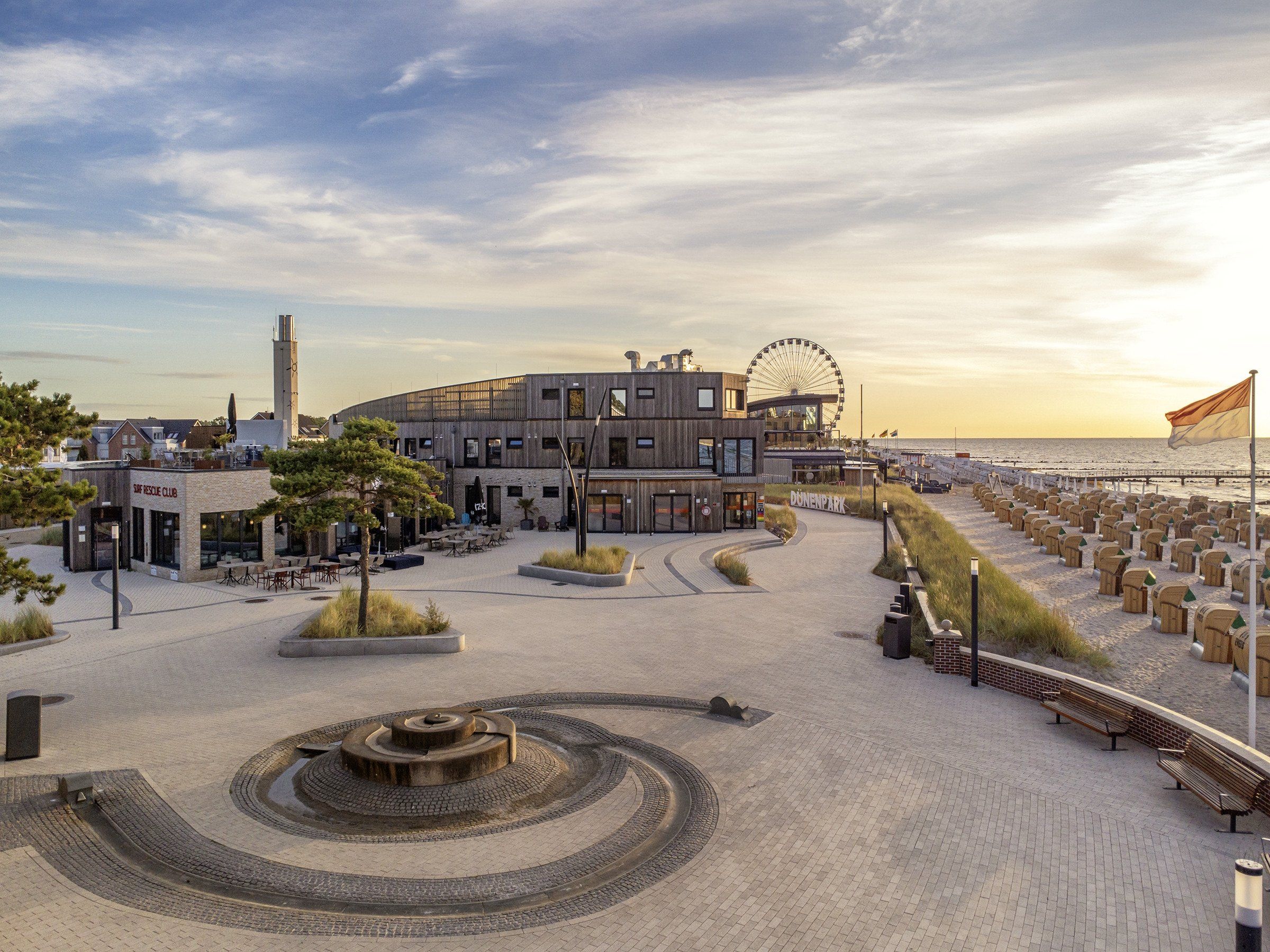 En havne-promenade med en moderne bygning, et pariserhjul og en strand med træhytter.