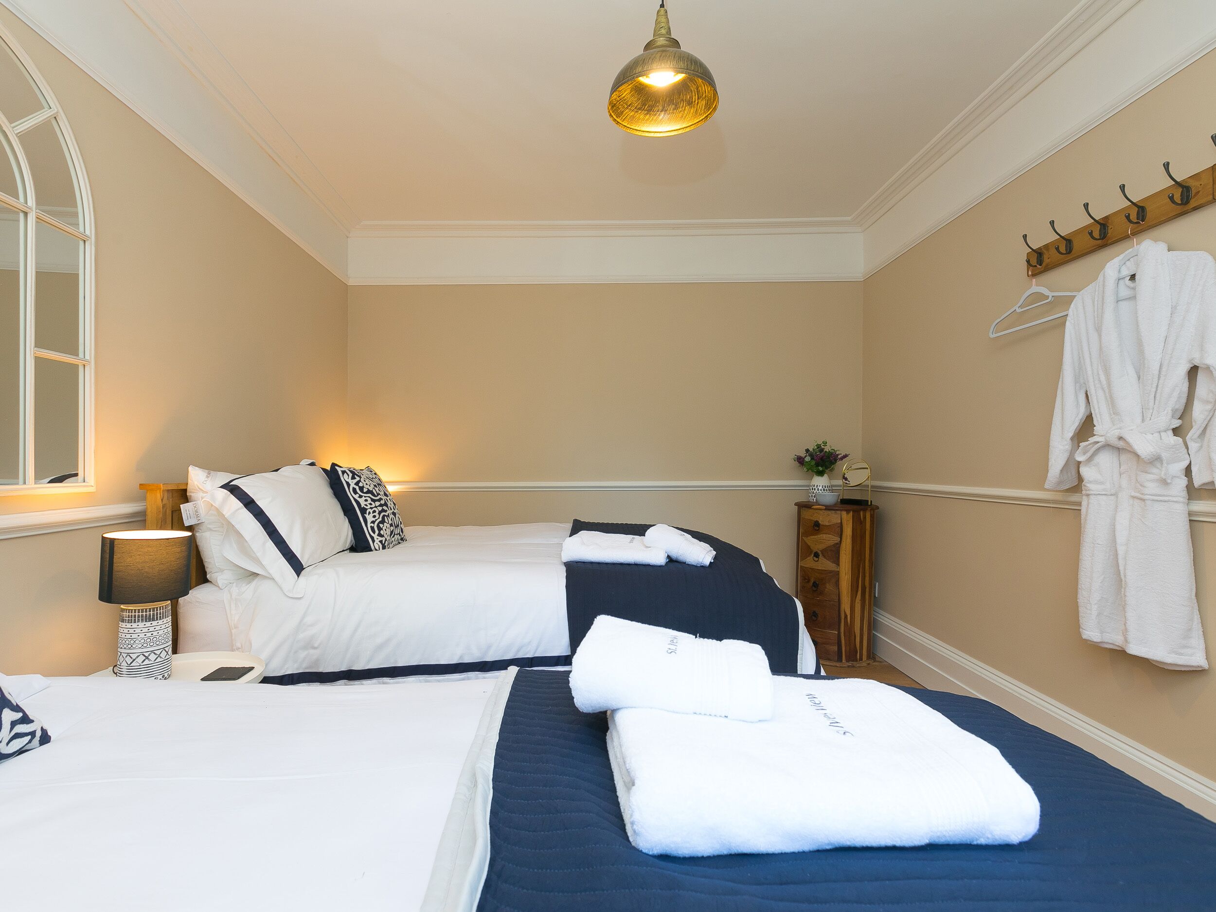 A hotel room with two beds, a bedside table with a lamp, a mirror, a wooden sideboard, and a robe hanging on a hook.