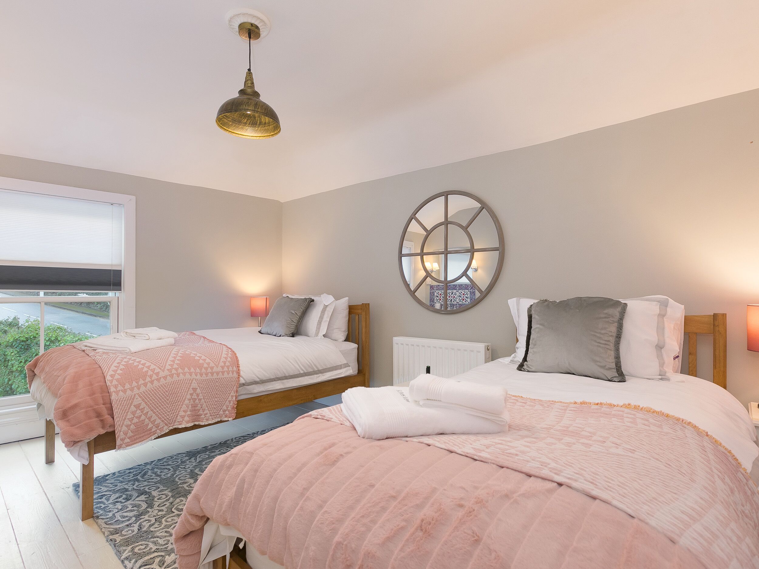 A bedroom with two beds, a large window, a ceiling light, and a wall-mounted mirror.
