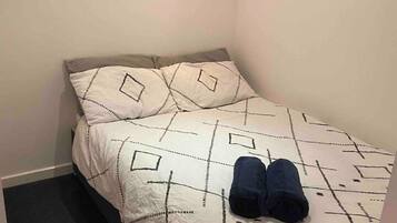 Una cama pequeña y bien hecha con una colcha y almohadas de estampado geométrico, un edredón oscuro doblado al pie de la cama, y una pared lisa con un interruptor de luz.