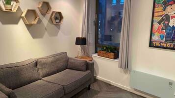 Una sala de estar moderna con un sofá gris, estanterías hexagonales de madera, una lámpara de pie y una ventana con cortinas.