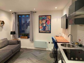 Un appartement moderne avec un canapé gris, un coin repas avec une chaise bleue, une cuisine équipée d’un évier et d’une cuisinière, et une fenêtre avec des rideaux.