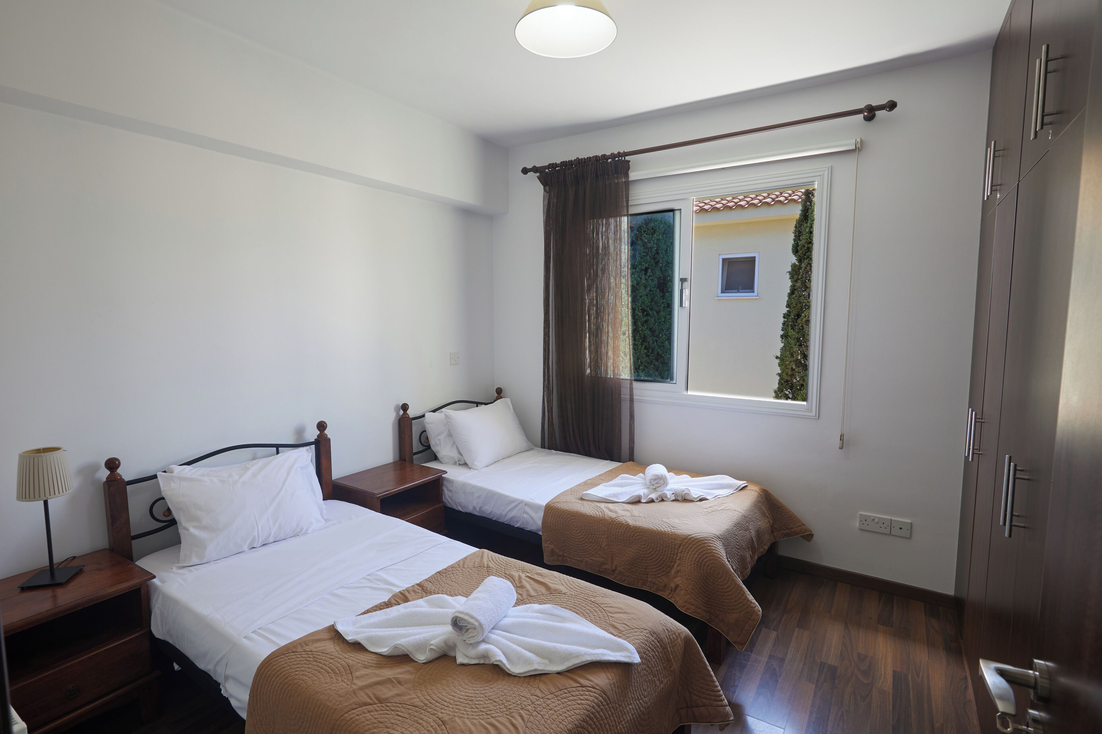 A hotel room with two beds, a window with curtains, a bedside table with a lamp, and a wardrobe.