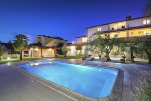Une piscine dotée d’une plage en pierre, entourée d’un jardin bien entretenu et d’une grande maison avec de nombreux balcons.