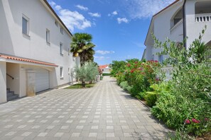 Pátio pavimentado com palmeira e plantas floridas, cercado por edifícios brancos com varandas.
