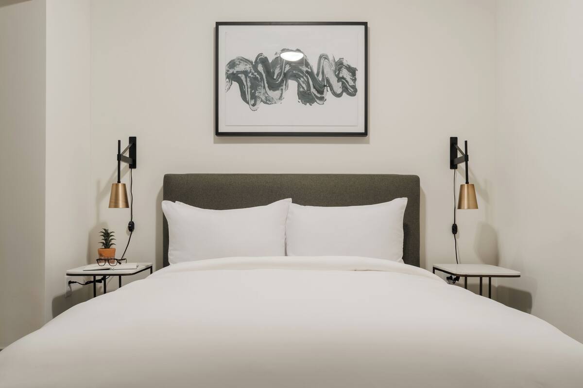 Un lit bien fait, avec du linge de lit blanc, une tête de lit grise et une œuvre d’art encadrée au-dessus. Deux tables de chevet avec des lampes sont placées de chaque côté du lit.
