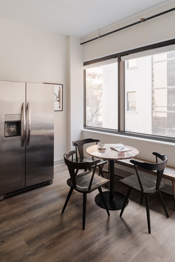Une cuisine moderne dotée d’un réfrigérateur en acier inoxydable, d’une table de salle à manger ronde avec quatre chaises et d’une grande fenêtre offrant une vue sur l’extérieur.