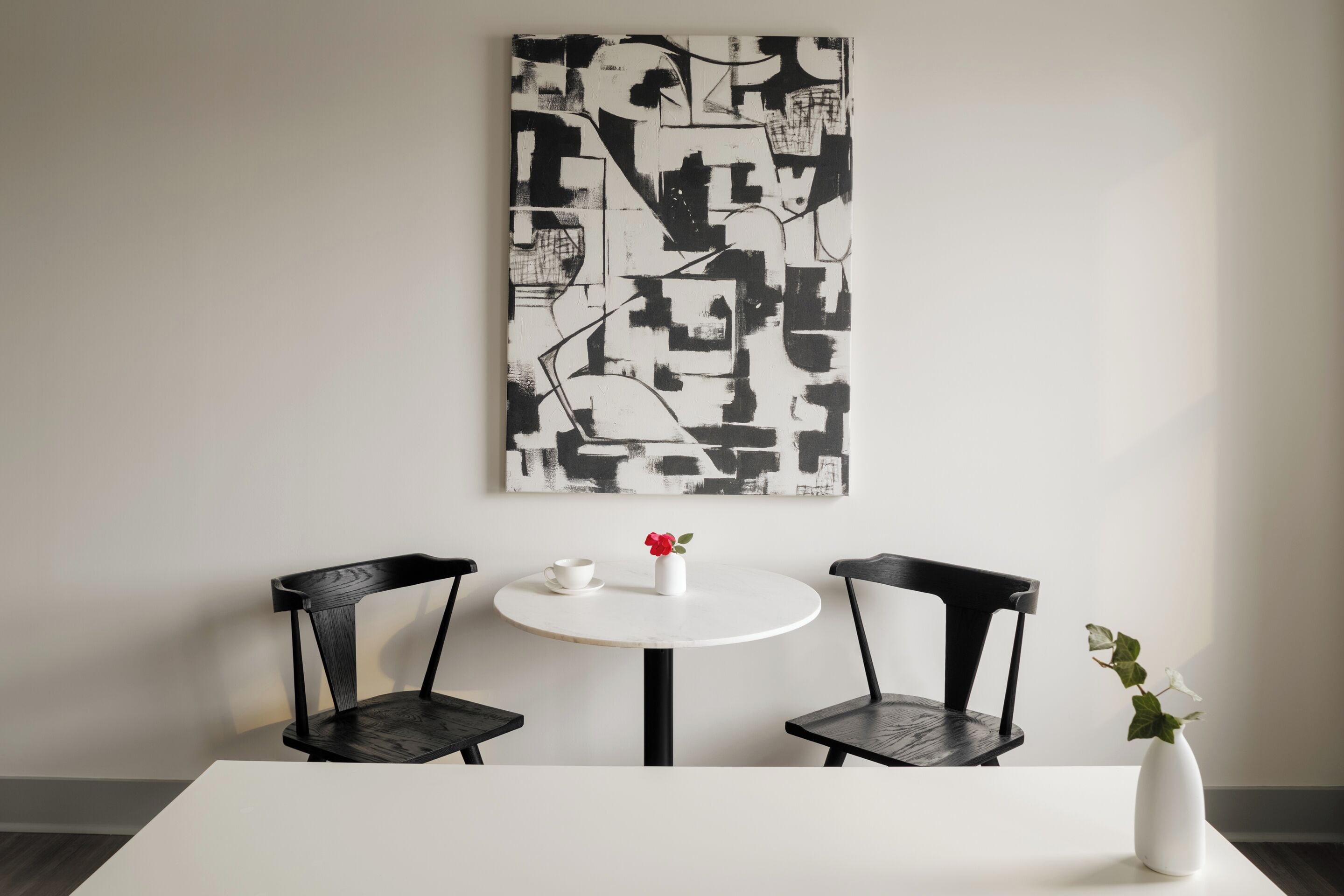 A minimalist room with a white table, two black chairs, a white vase with a red flower, and a black and white abstract painting on the wall.