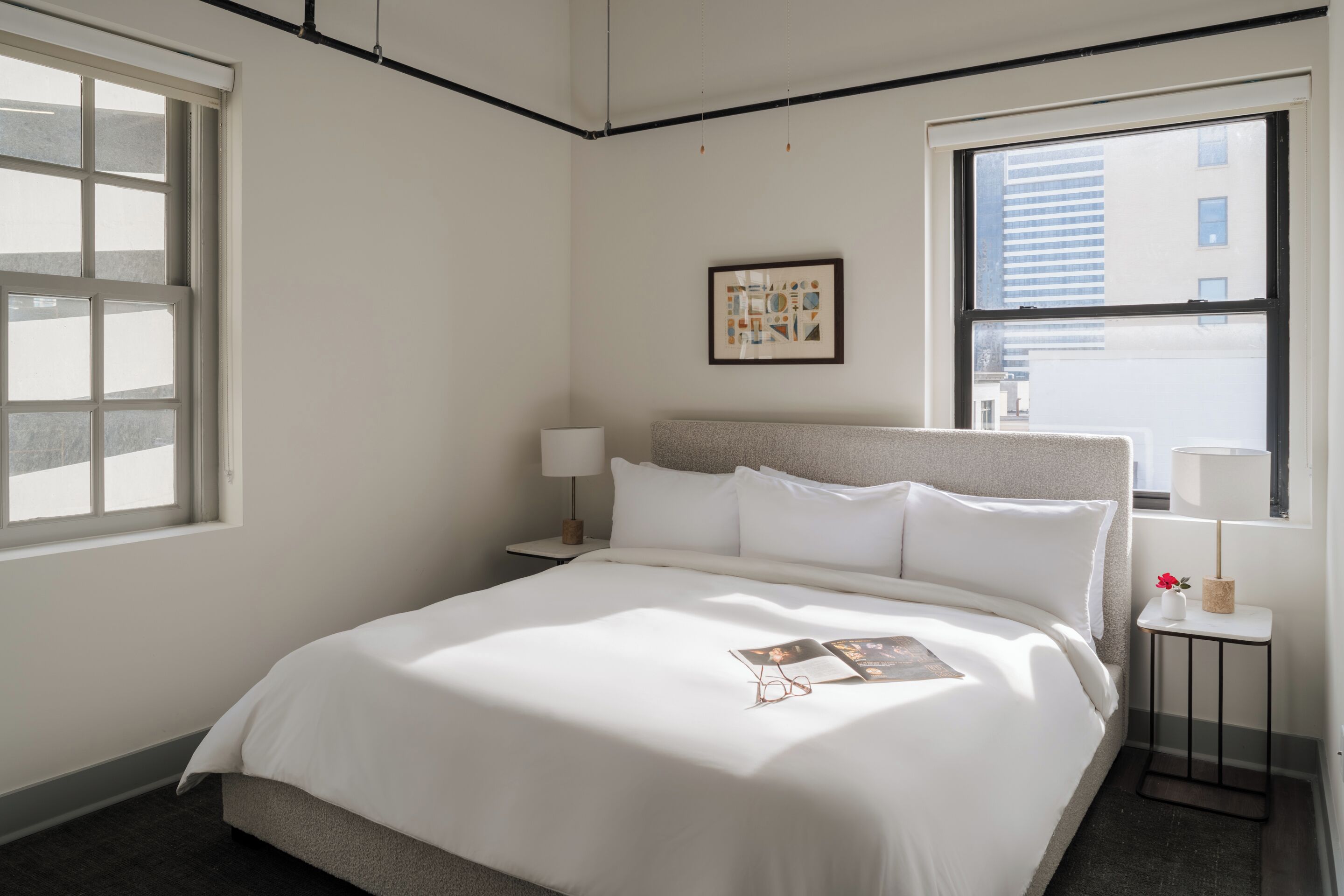 A hotel room with a large bed, a bedside table with a lamp, a small table with a vase and a picture frame, and a window with a view of buildings.