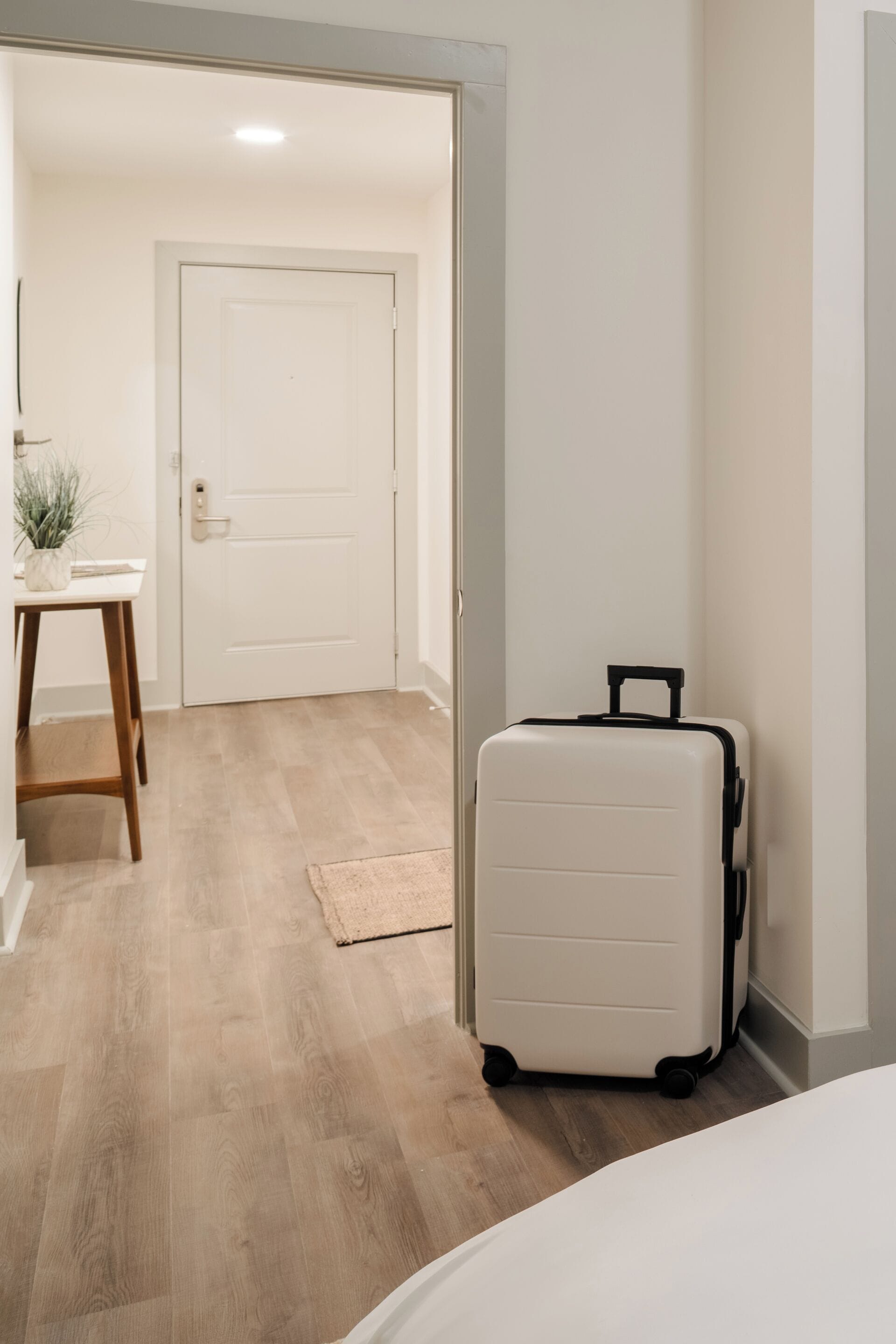 Habitación de hotel con suelo de madera, cama blanca, maleta blanca y puerta que da a otra habitación.