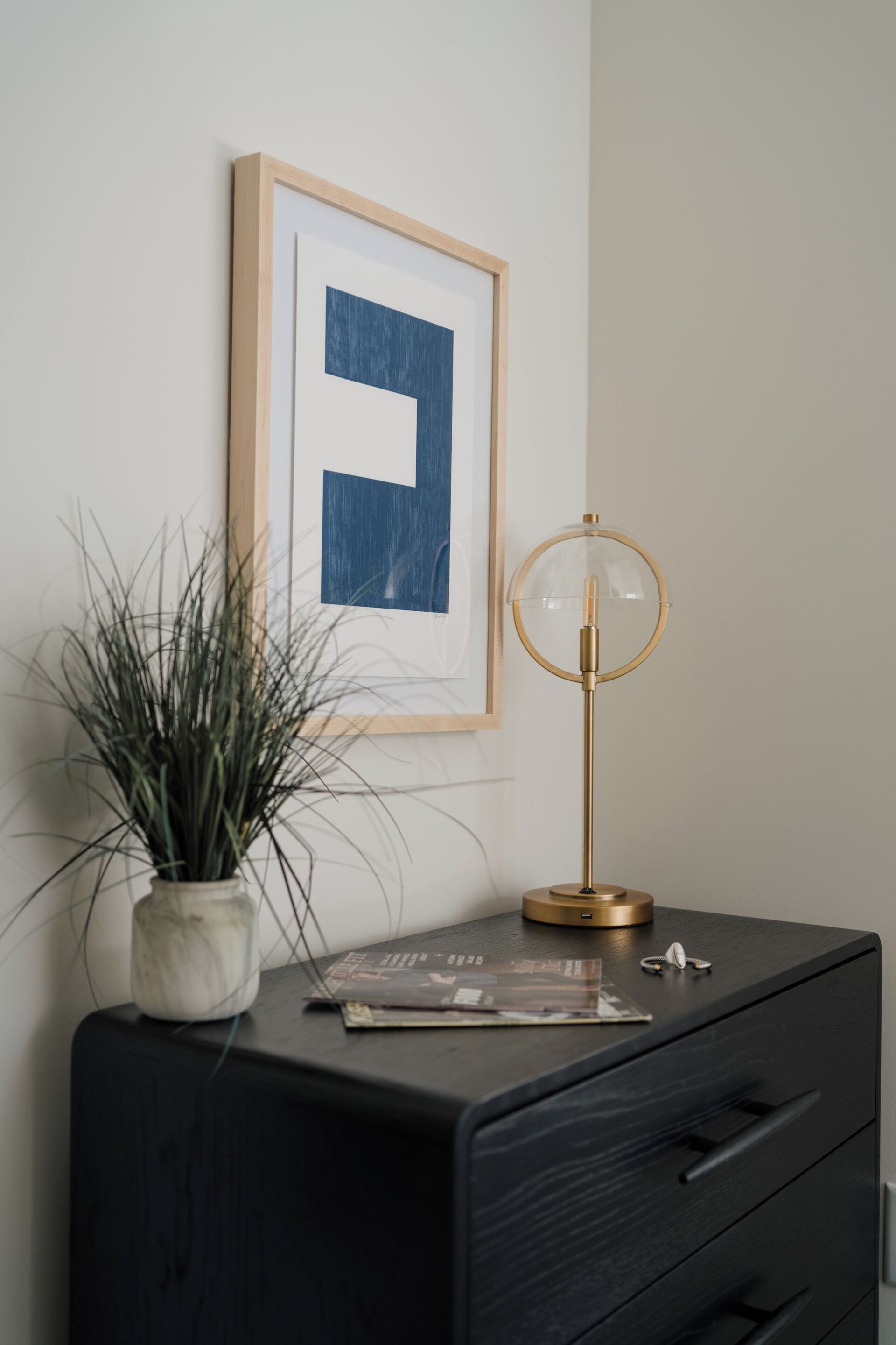 Une table de chevet noire ornée d’un tableau encadré, d’une plante en pot, d’une lampe et d’un magazine.
