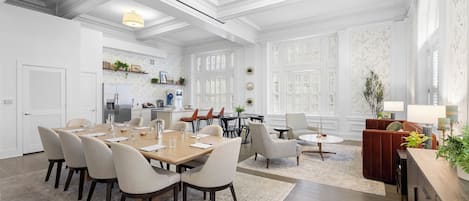 Un vaste espace de salle à manger avec une longue table en bois et de nombreuses chaises, un coin salon confortable avec un canapé rouge et des fauteuils, et une cuisine équipée d’appareils modernes.
