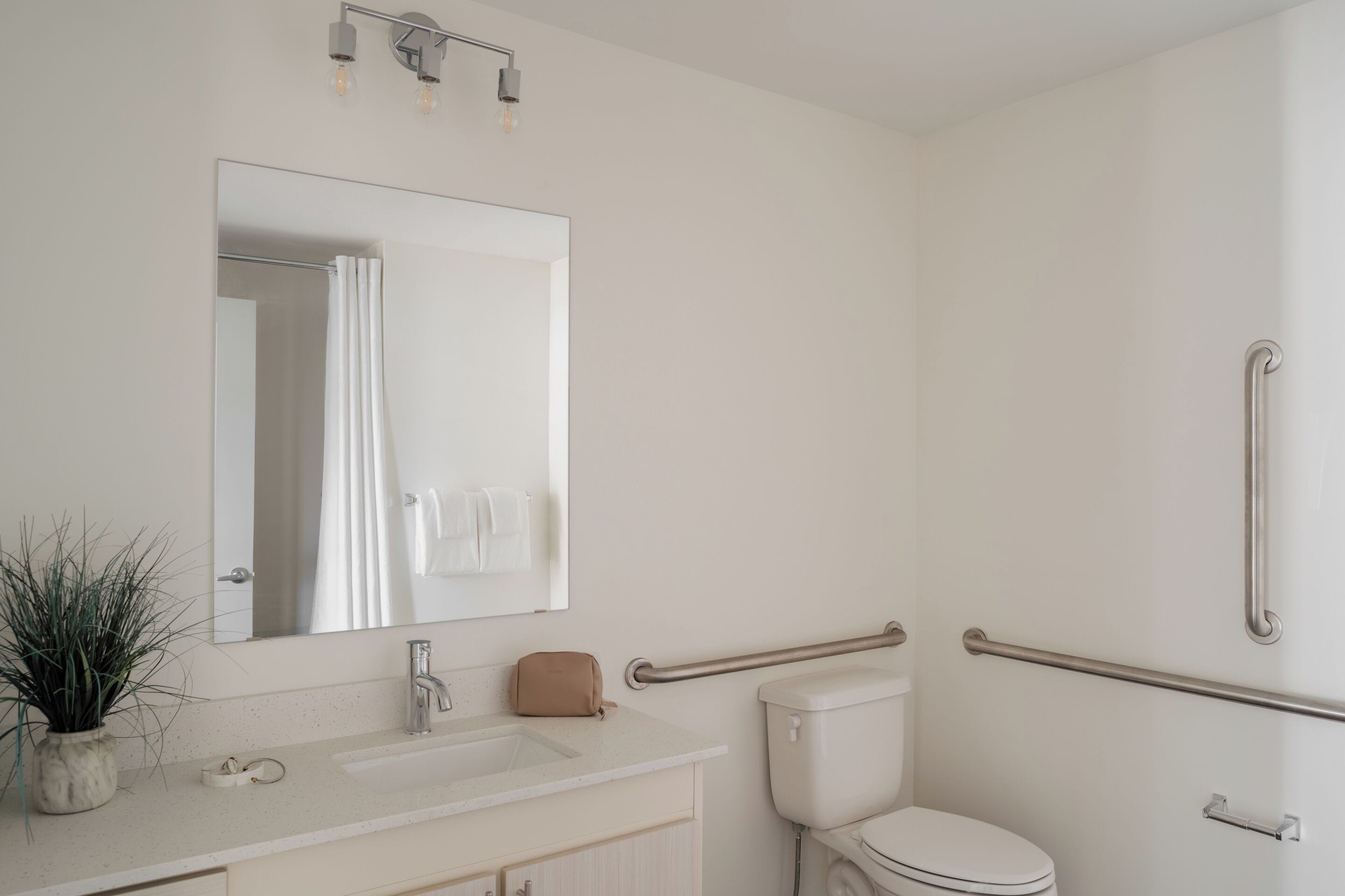 A modern bathroom with a white sink, mirror, and toilet. There are handrails on the walls for accessibility.