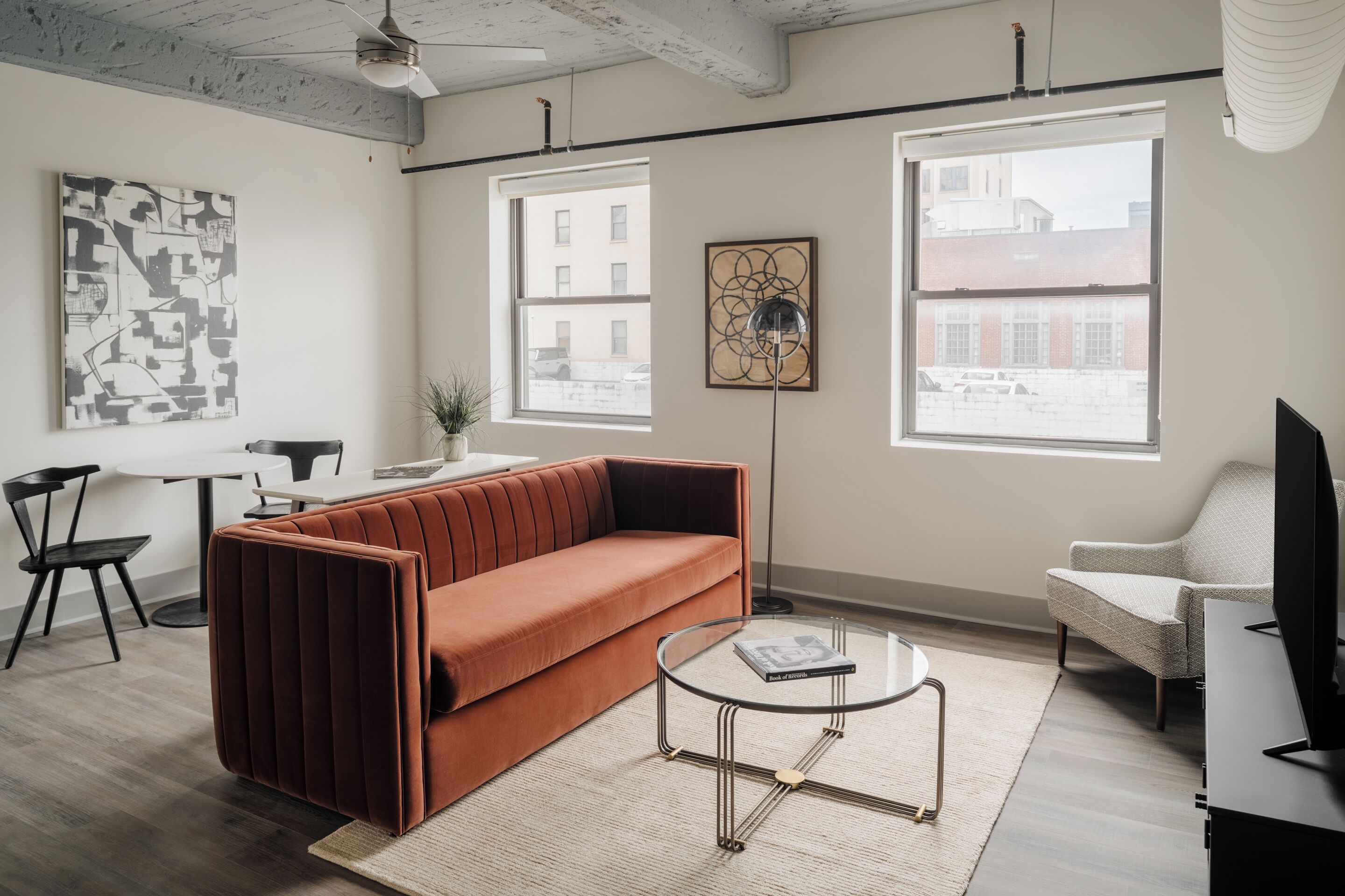 A modern living room with a brown sofa, a glass coffee table, and a white armchair. There are two windows providing natural light and a view of buildings outside.
