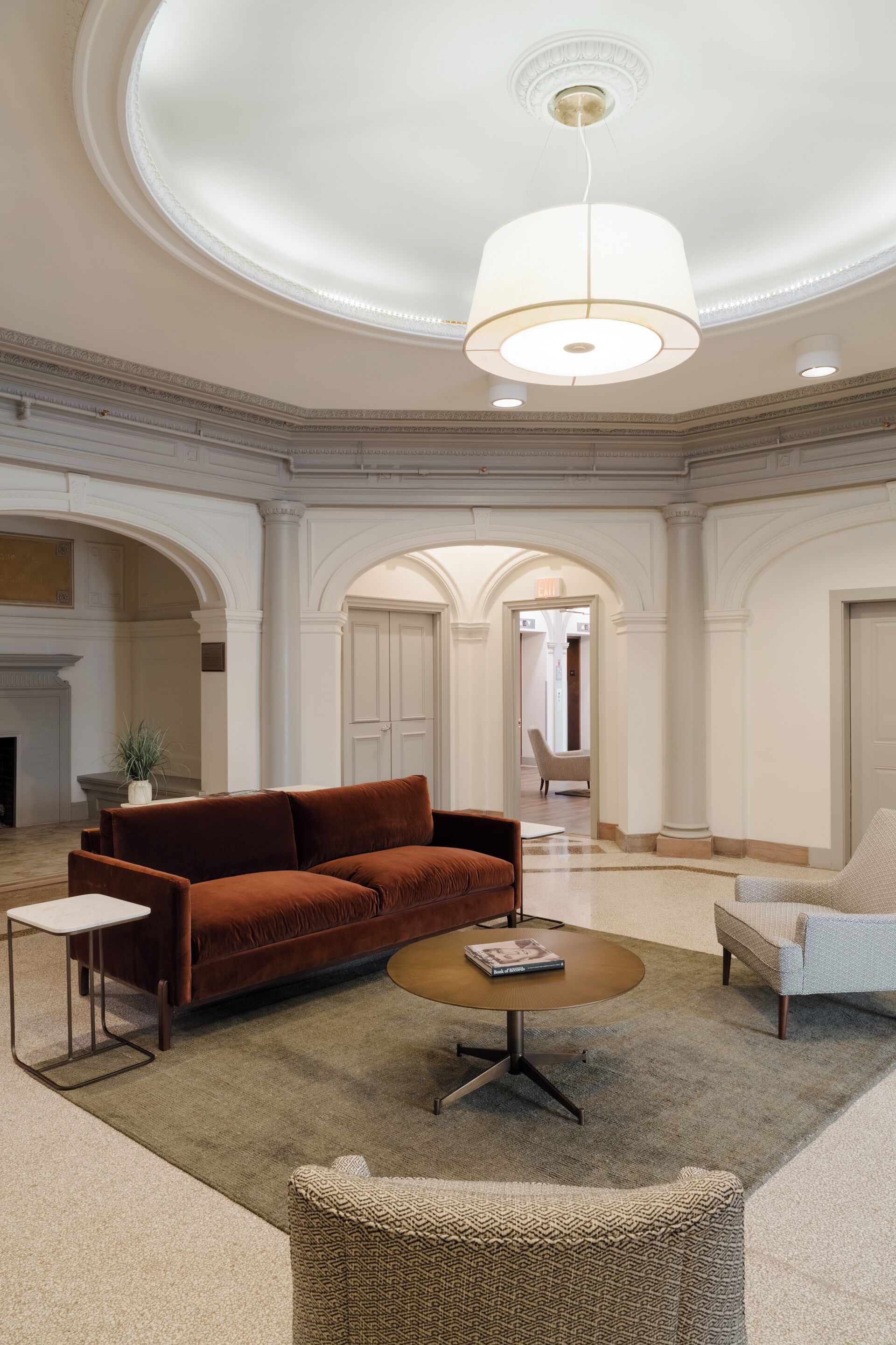 A spacious lobby with a large brown sofa, a round coffee table, and a white armchair. There is a fireplace and a plant in the corner.
