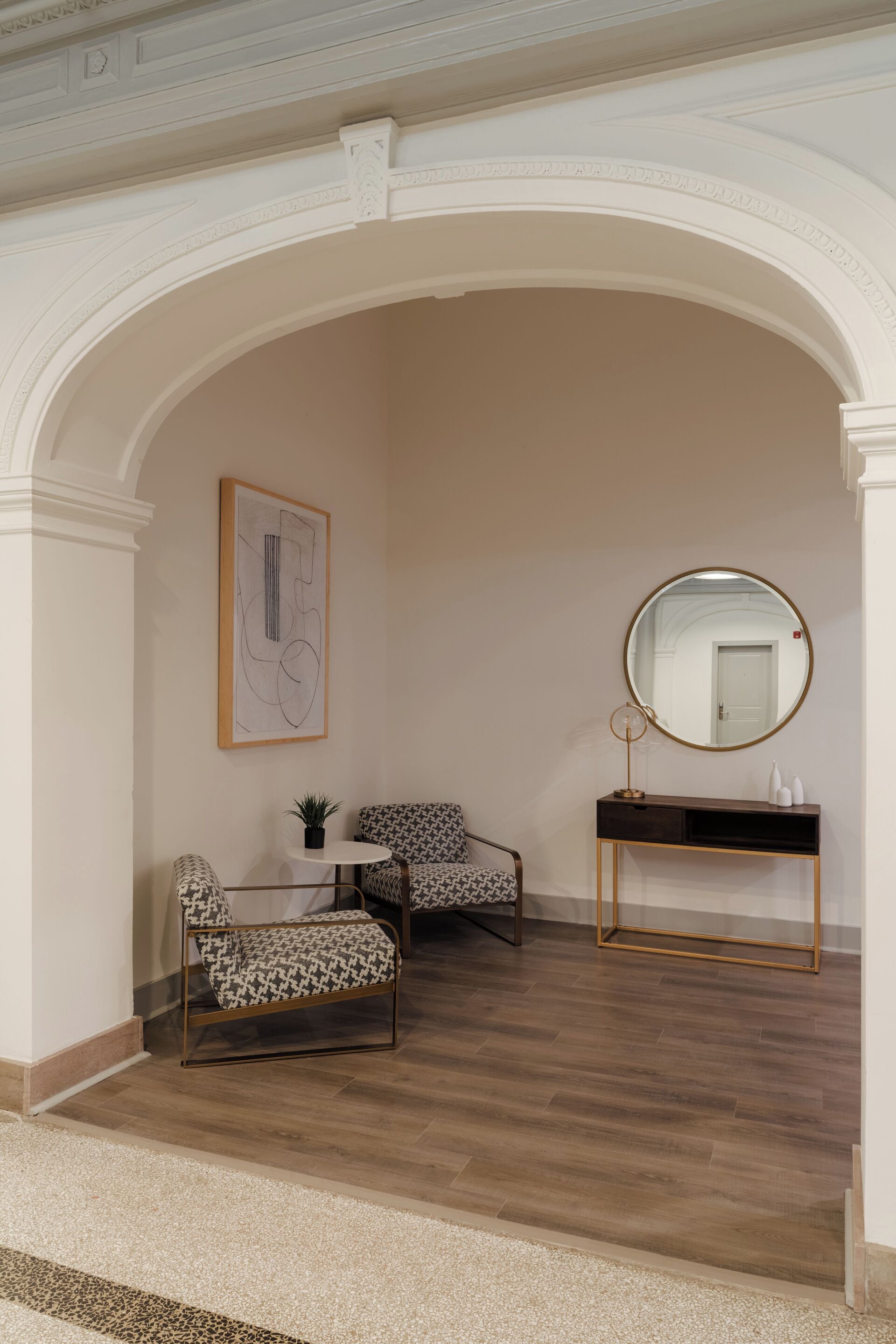 A room with a wooden floor, a round mirror on a console table, two patterned chairs, a small table with a plant, and a framed artwork on the wall.