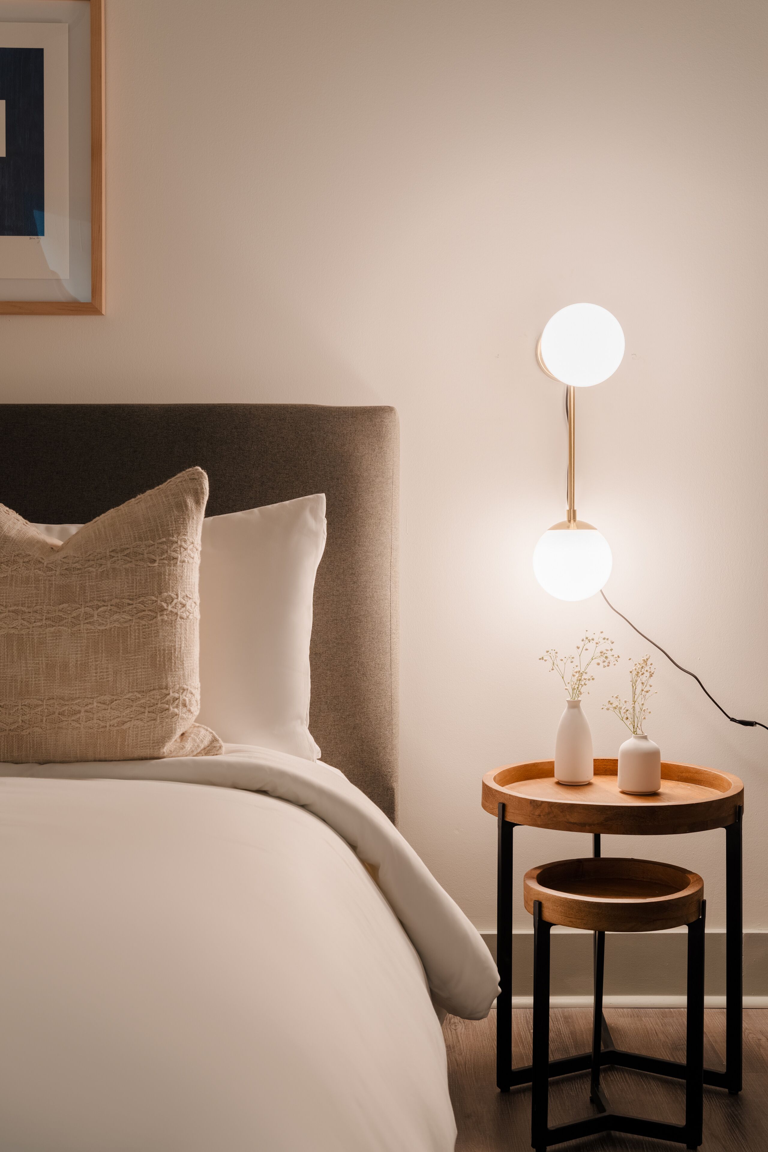 Een net opgemaakt bed met hoofdbord, een nachtkastje met lamp en een vaas met bloemen.