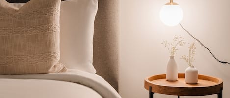 Ein ordentlich bezogenes Bett mit Kopfteil, ein Nachttisch mit Lampe und eine Vase mit Blumen.
