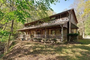 A rustic wooden cabin with a porch, surrounded by trees and a grassy area.