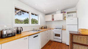 A modern kitchen with white cabinetry, a wooden countertop, a gas stove, a dishwasher, a microwave, and a refrigerator. - Springs Beach House - Point Lonsdale (Point Lonsdale)