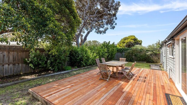 Una terraza de madera con mesa y sillas, rodeada por un cerco de madera y abundante vegetación.