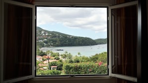 A window with a view of a coastal area, including a body of water, boats, and buildings on a hillside. - 3-bedroom apartment with panoramic view of Anse à l'Ane bay, 2 steps from the beach (Les Trois-Îlets)