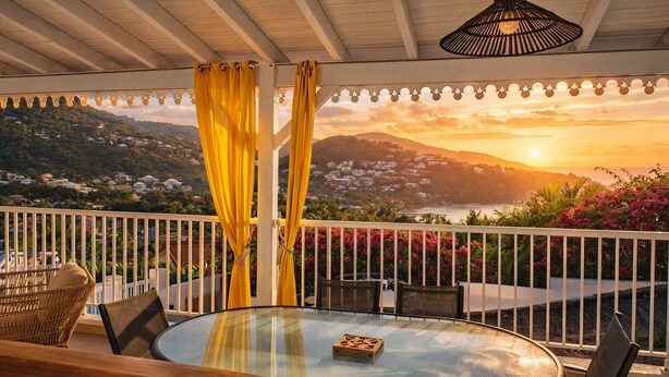 A balcony with a glass table, yellow curtains, wicker chairs, and a view of a sunset over a mountainous landscape. - 3-bedroom apartment with panoramic view of Anse à l'Ane bay, 2 steps from the beach (Les Trois-Îlets)