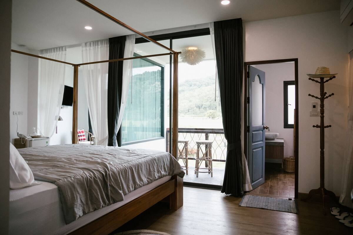 A bedroom with a large bed, a wooden floor, a balcony with a view, and a bathroom with a sink and mirror.