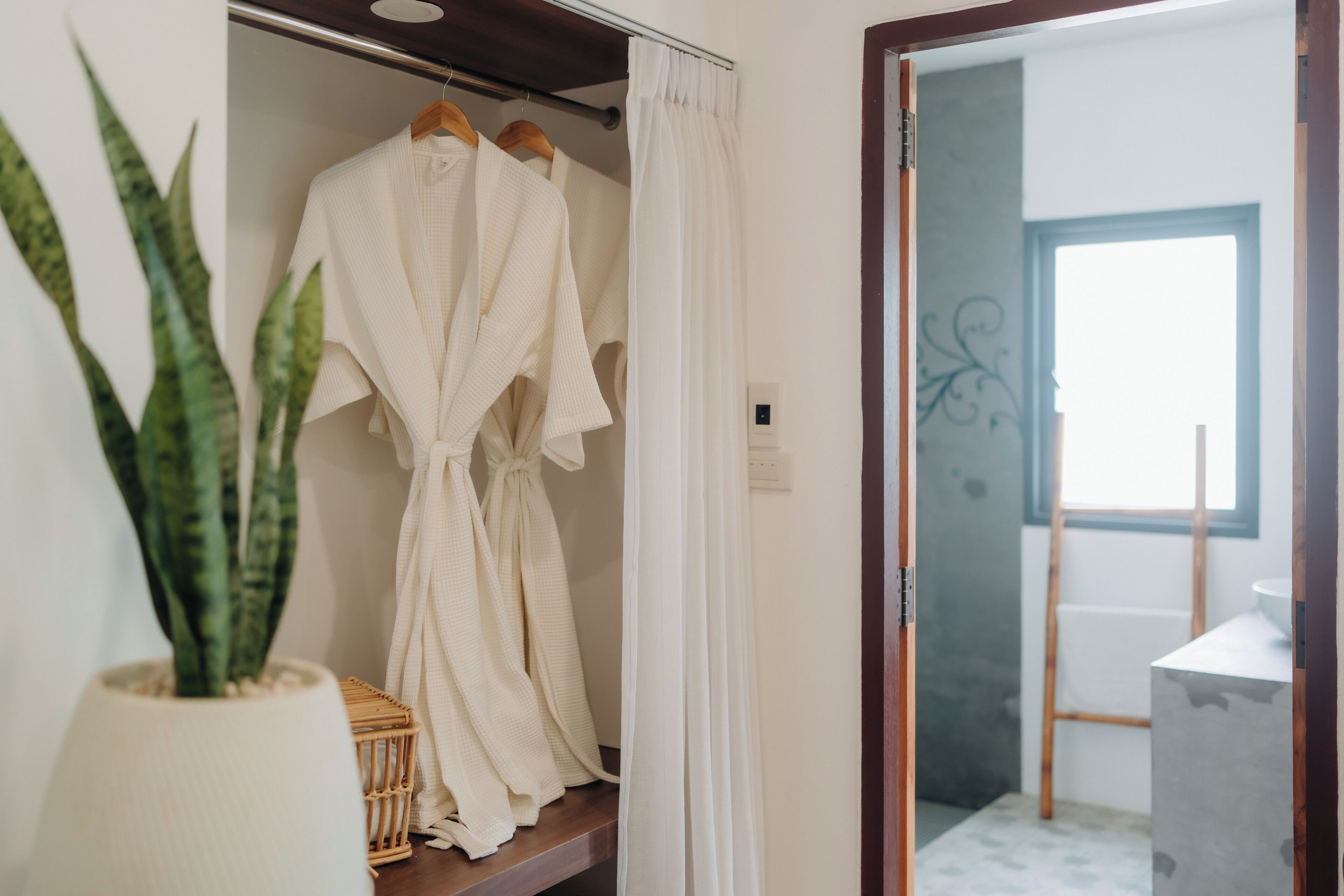 A room with a wardrobe containing two white robes, a potted plant, and a wooden shelf.