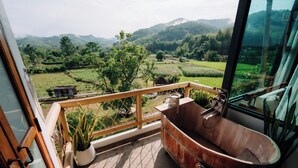 Un balcón con vistas a campos verdes y montañas, una bañera de madera y una planta en maceta.