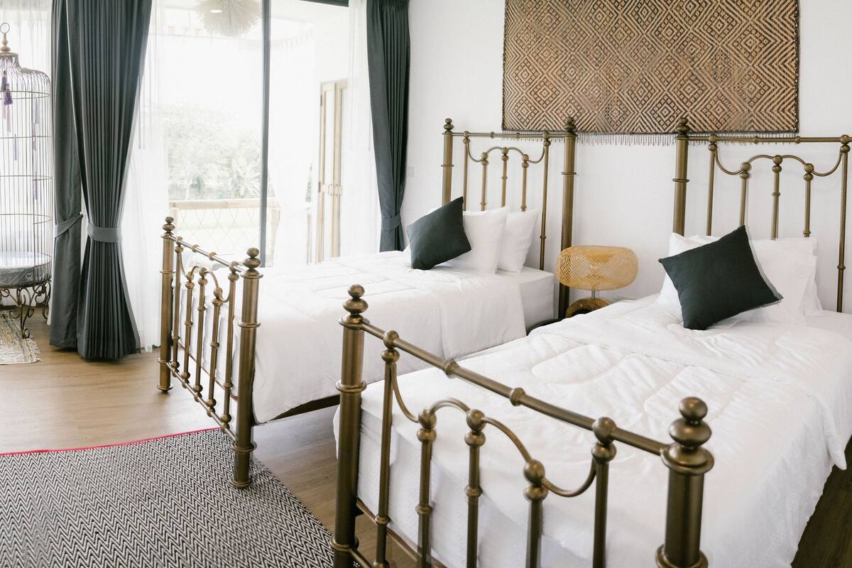 A bedroom with two beds, a window with curtains, a birdcage, and a patterned rug.