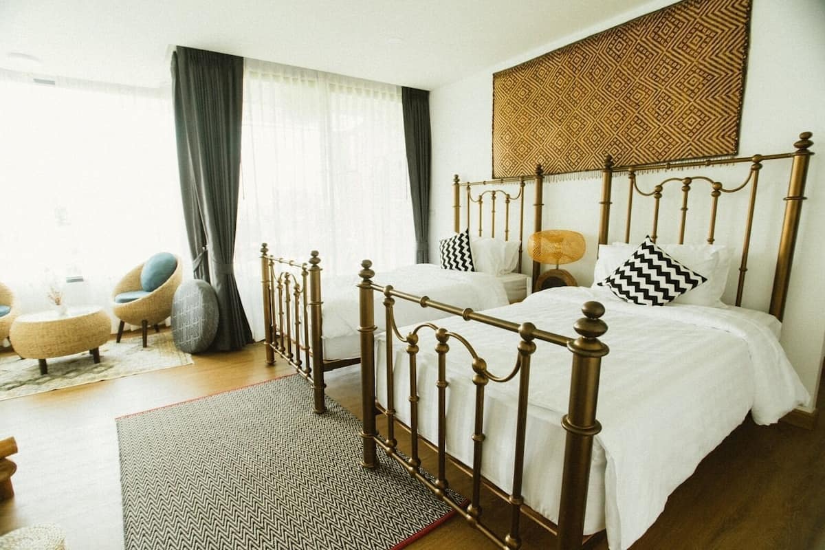 A bedroom with a wooden four-poster bed, a sitting area with chairs, a patterned rug, and a large window with curtains.