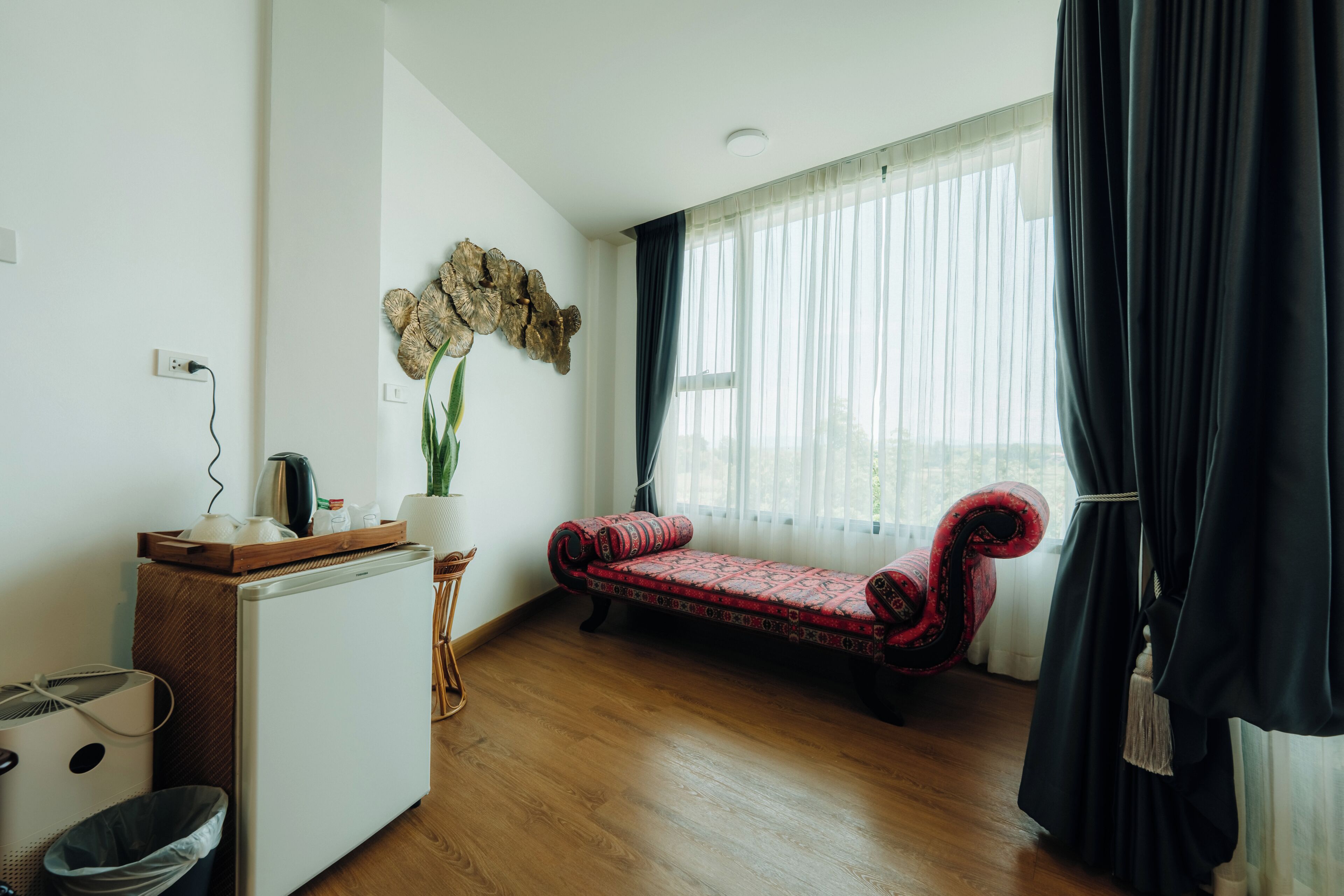 A room with a wooden floor, a white refrigerator, a red patterned sofa, a potted plant, and a large window with sheer curtains.