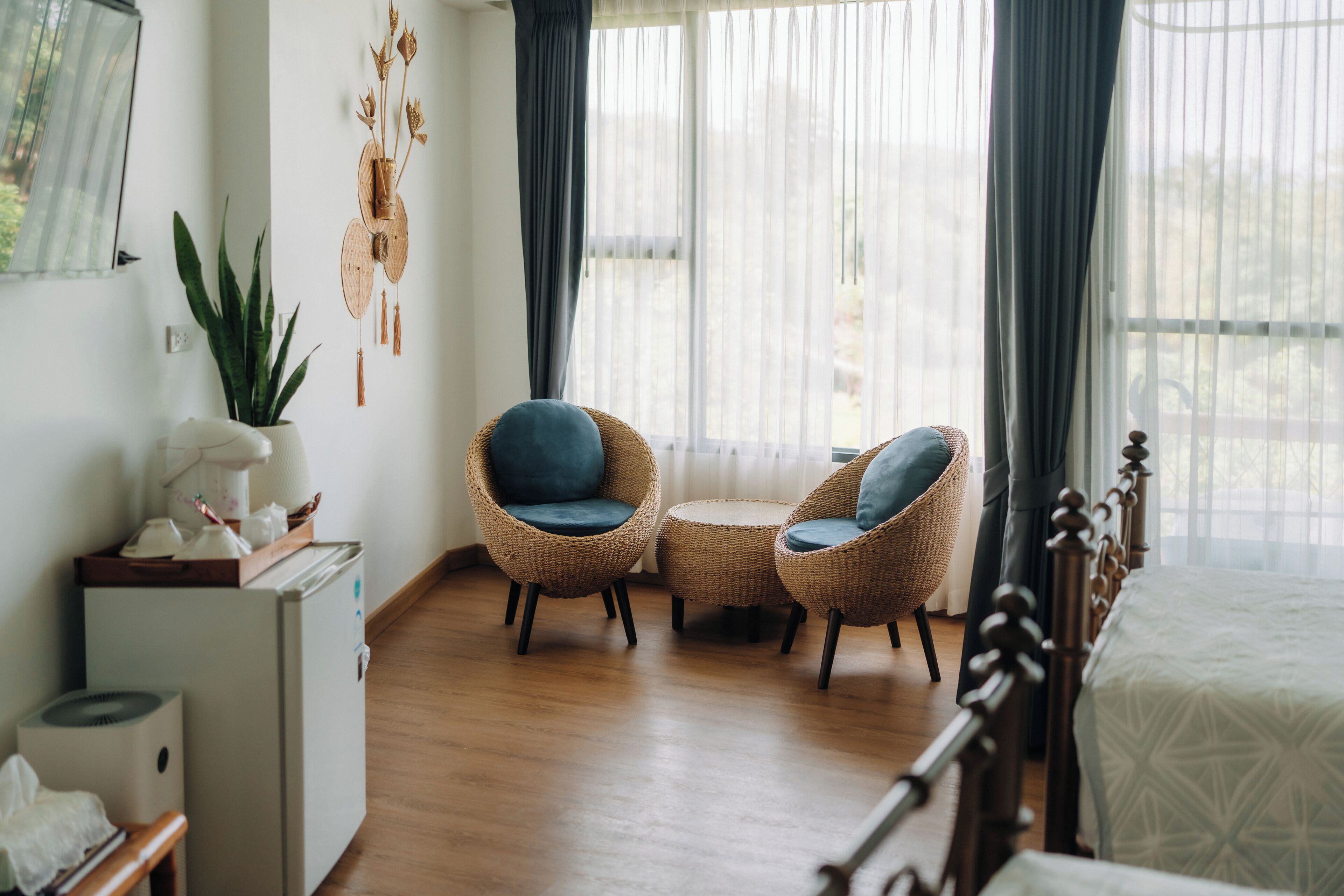 A room with a bed, two wicker chairs with blue cushions, a small table, a white desk with a lamp, a potted plant, and a wall decoration.