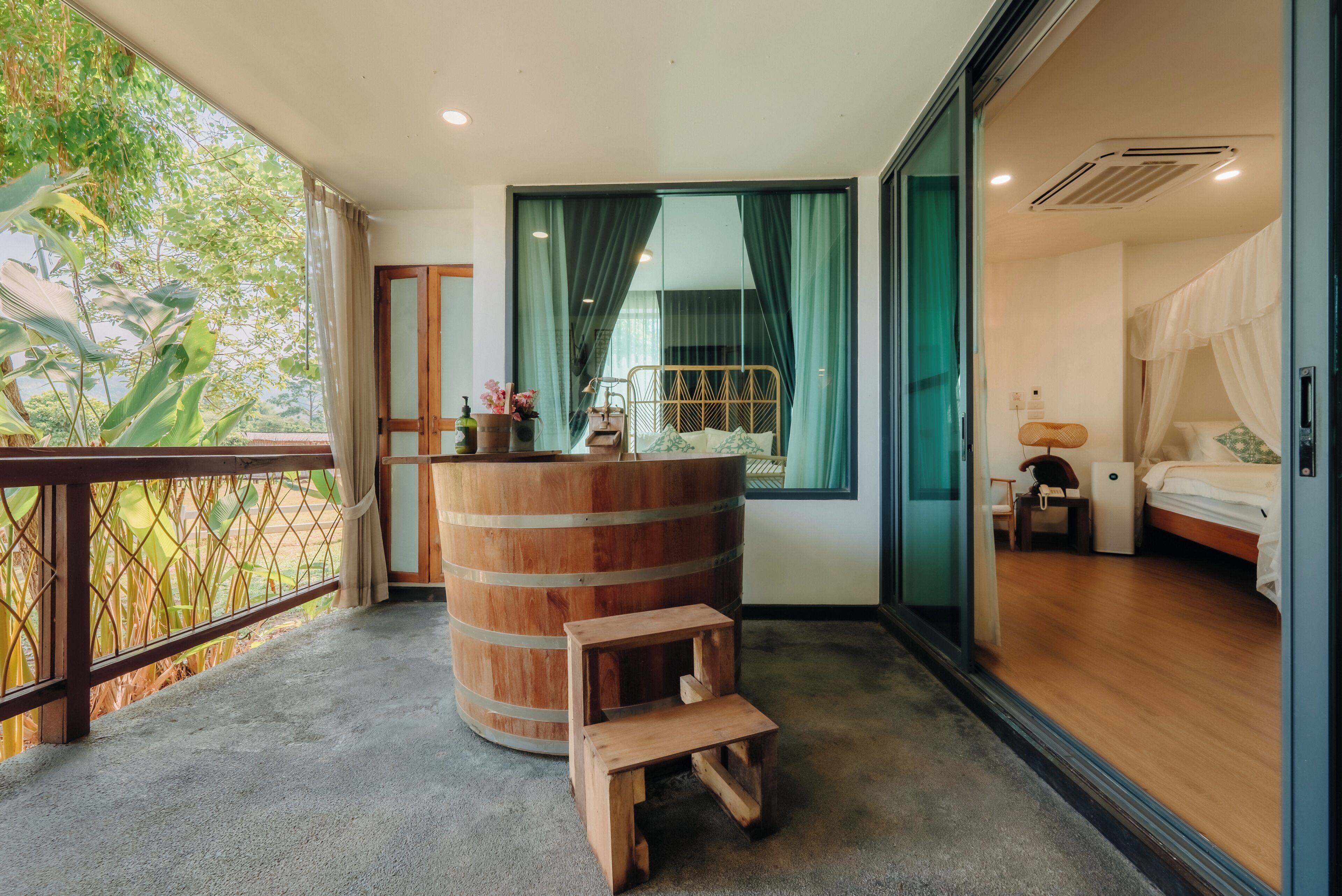 Une pièce comprenant un lit, un petit tabouret en bois, une grande barrique en bois et une porte-fenêtre coulissante donnant sur un balcon avec vue sur la verdure.