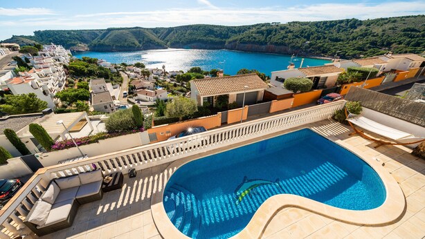 Una piscina con bordo curvo, circondata da lettini e un ombrellone, con vista su una cittadina costiera e case a terrazza e vista sul mare.