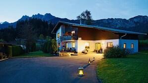 A two-story chalet with a balcony, a driveway, and a mountain backdrop.