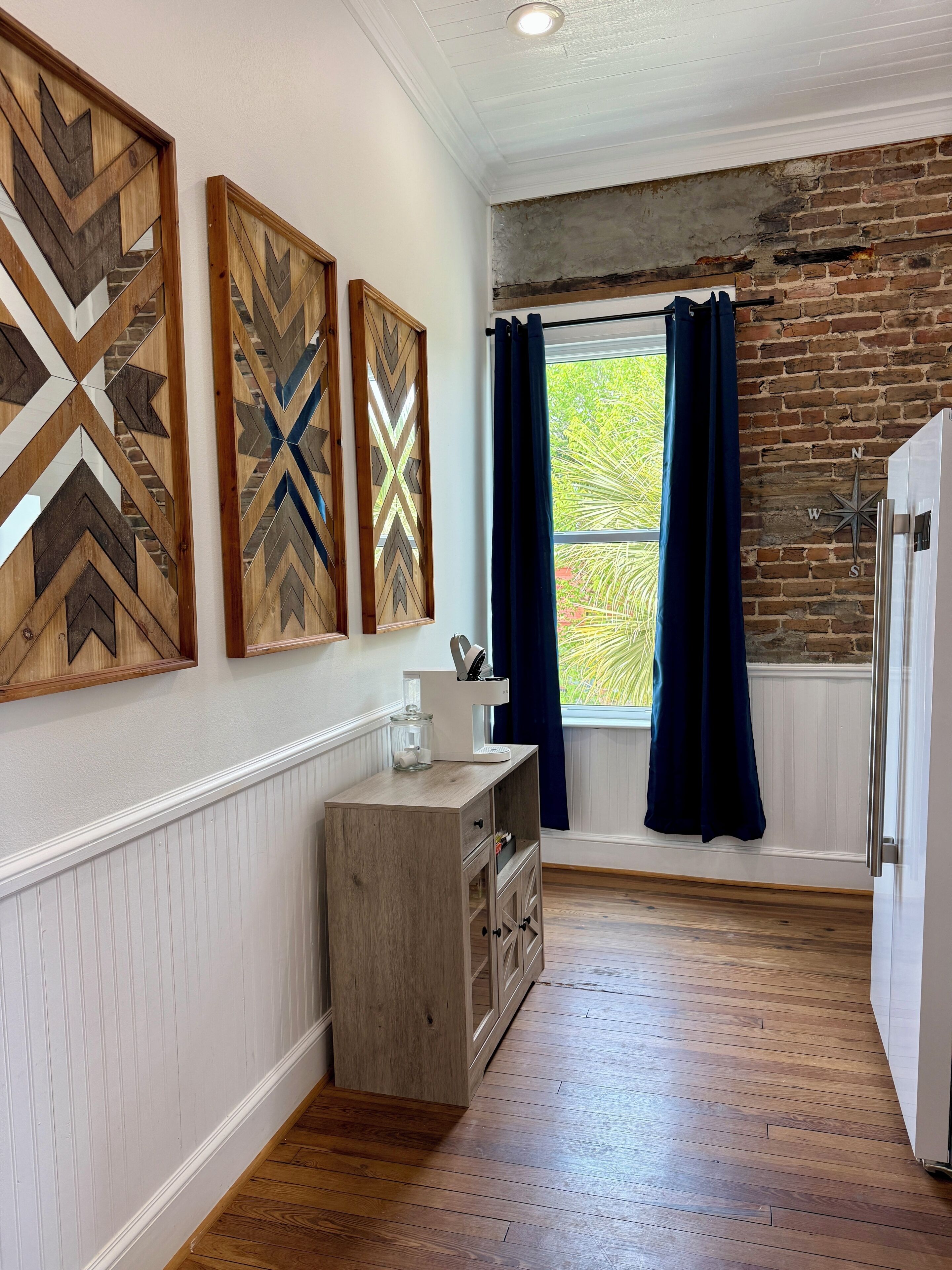 Una cocina con piso de madera, pared de ladrillo, ventana con cortinas y un refrigerador.