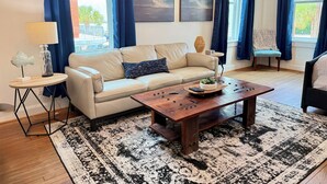 A living room with a beige sofa, a wooden coffee table, a patterned rug, and a view of the outdoors through large windows with blue curtains. - In the heart of downtown Apalach w/ restaurants, shops & waterfront within steps (Apalachicola)