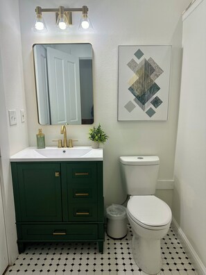 Un baño con lavamanos blanco, tocador verde, grifos dorados, inodoro blanco y una obra de arte con motivos geométricos en la pared.