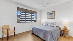 Une chambre avec un grand lit, une chaise, une fenêtre avec des stores, un ventilateur de plafond et un tableau encadré au mur.