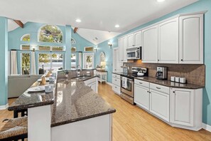 A kitchen with white cabinets, a granite countertop, and a view of the outdoors through large windows.
