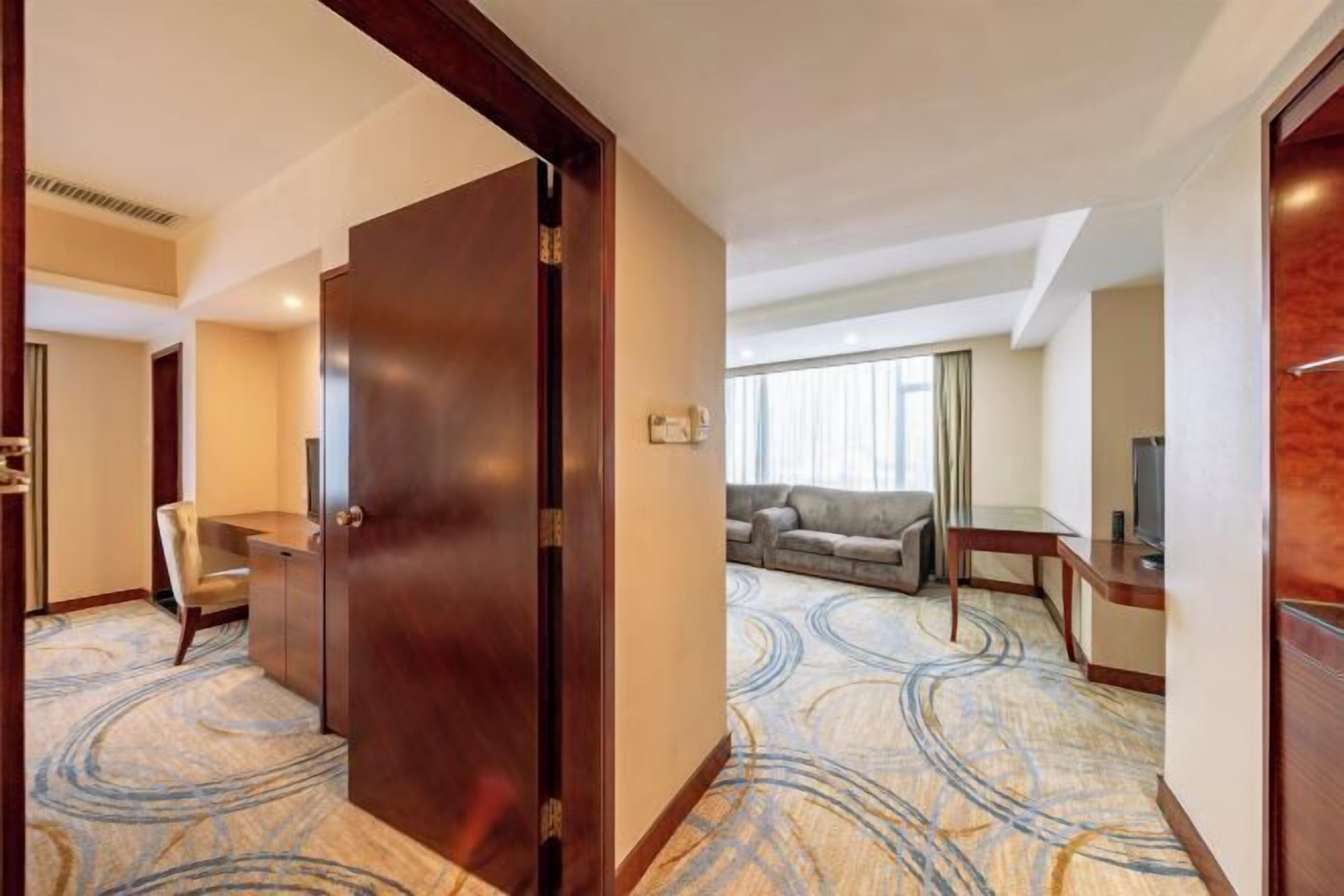 A hotel room with a large wooden door, a seating area with a sofa and armchairs, a small table, and a television.